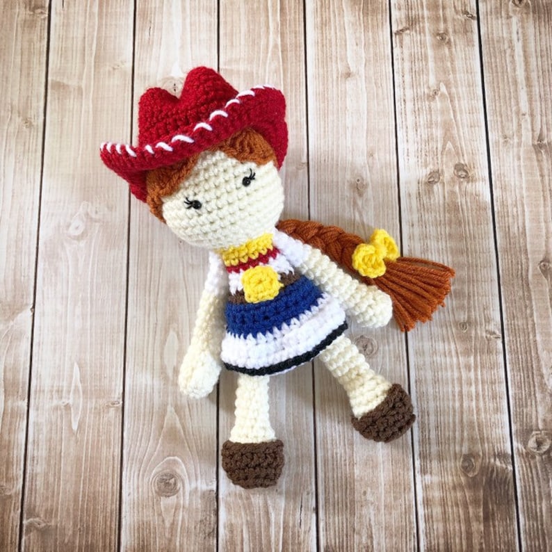 May include: Crocheted doll with a red and white cowboy hat, a white dress with blue and yellow accents, and brown hair. The doll is lying on a wooden surface.