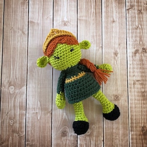 May include: Green crocheted doll with a brown and yellow crown, green dress, and black shoes. The doll has a long orange braid.