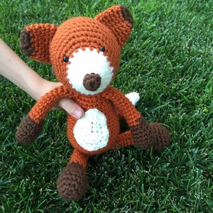 May include: A crocheted fox plush toy with brown and orange fur, white accents, and black eyes. The toy is sitting on a bed of green grass.