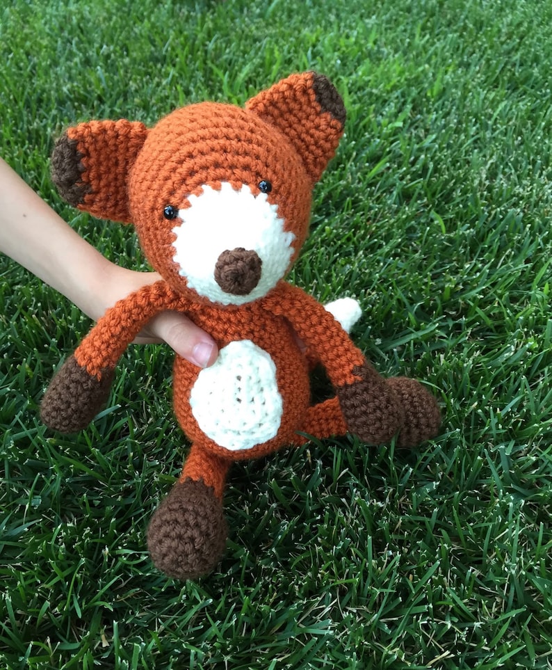 May include: A crocheted fox plush toy with brown and orange fur, white accents, and black eyes. The toy is sitting on a bed of green grass.