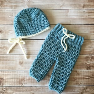 May include: A blue crocheted baby hat and trousers set with a white crocheted trim. The hat has a white tie.