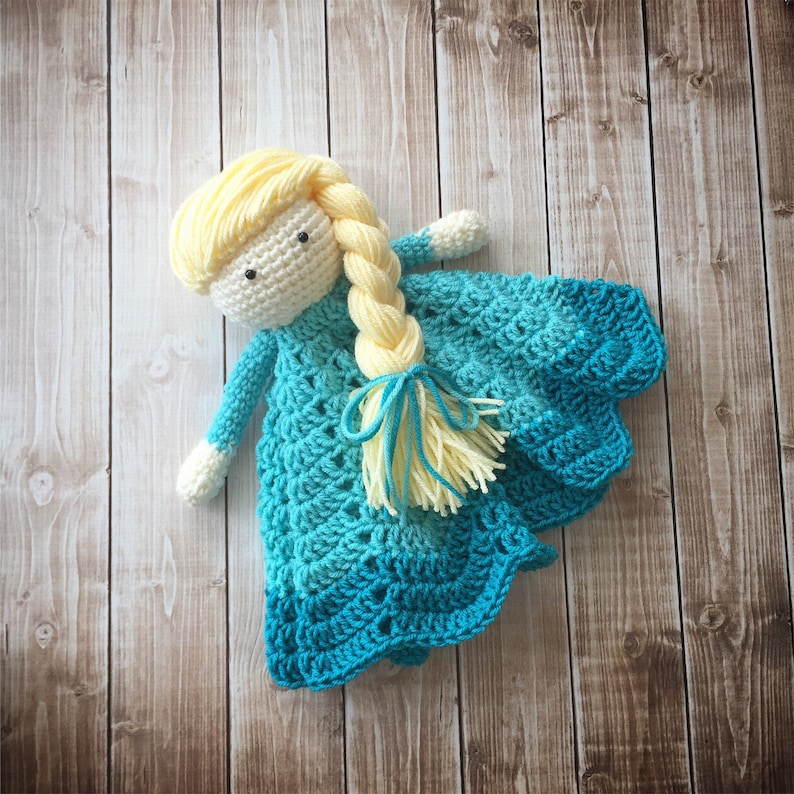 May include: A crocheted doll with a blue and green dress and long blonde hair. The doll is lying on a wooden surface.