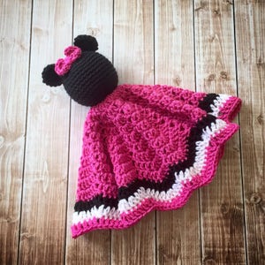 May include: A pink crocheted baby blanket with a black and white trim. The blanket has a black crocheted mouse head attached to the top.
