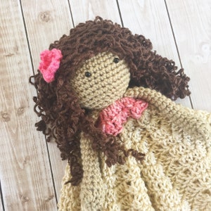 Pode incluir: Uma boneca de croch&ecirc; com cabelo castanho encaracolado e uma flor rosa. A boneca est&aacute; vestindo um vestido de croch&ecirc; bege com uma flor de croch&ecirc; rosa no ombro.