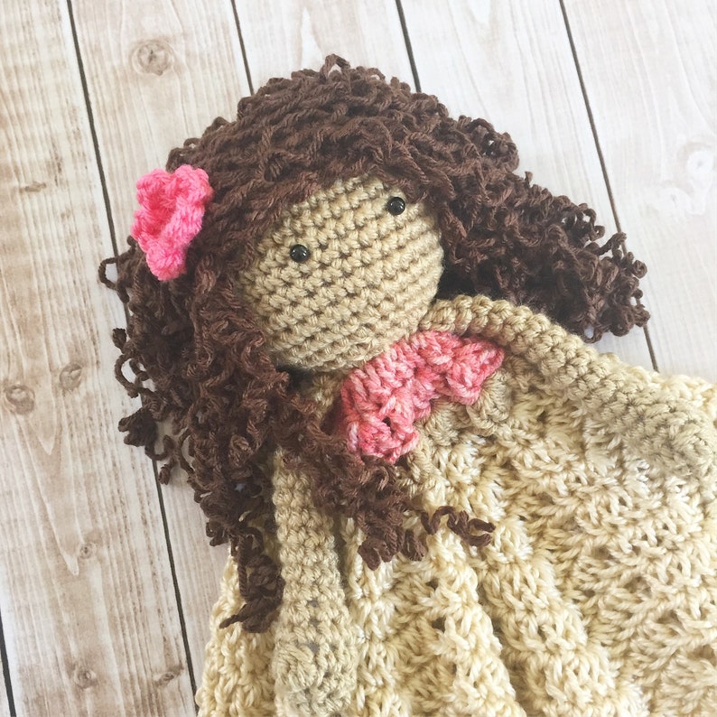 Pode incluir: Uma boneca de croch&ecirc; com cabelo castanho encaracolado e uma flor rosa. A boneca est&aacute; vestindo um vestido de croch&ecirc; bege com uma flor de croch&ecirc; rosa no ombro.