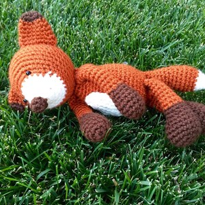 May include: A crocheted fox plush toy with brown and orange fur, white accents, and black eyes. The toy is lying on a bed of green grass.
