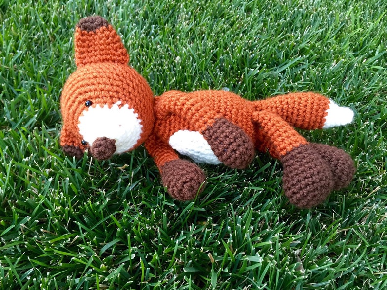 May include: A crocheted fox plush toy with brown and orange fur, white accents, and black eyes. The toy is lying on a bed of green grass.
