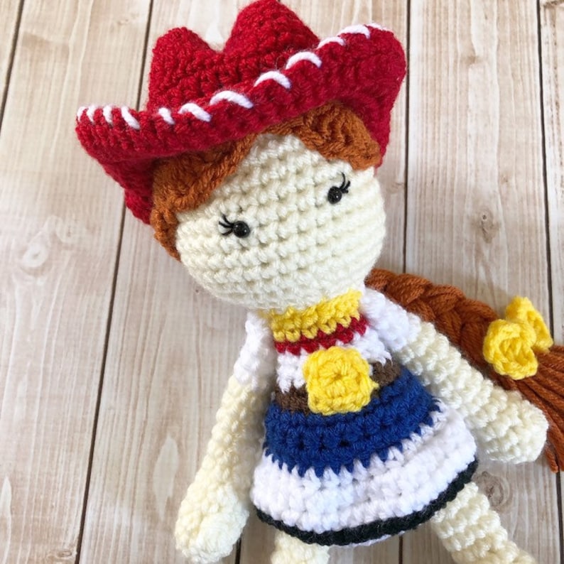 May include: Crocheted doll with brown hair, wearing a red and white cowboy hat, a white shirt with a yellow star, a blue and brown vest, and a blue skirt.