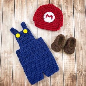 May include: A blue crocheted overall with yellow buttons, a red crocheted hat with a white 'M' on it, and two brown crocheted booties.