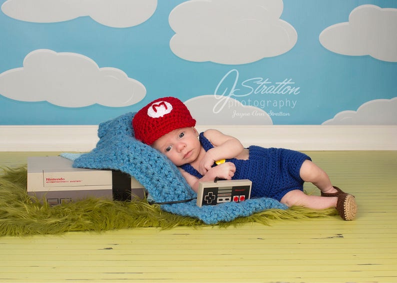 May include: A baby wearing a red and white crocheted Mario hat and blue overalls is lying on a blue blanket with a Nintendo Entertainment System console and a controller. The background is a blue and white cloudy sky.