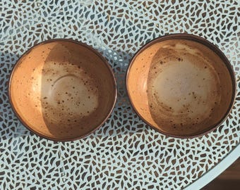 May include: Two round ceramic bowls with a light tan interior featuring dark brown speckles and a smooth, rounded rim. The bowls have a warm, earthy tone and a consistent circular shape, showcasing a simple design with a speckled finish.