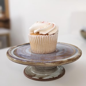May include: A single cupcake with white frosting and pink sprinkles sits on a ceramic cake stand. The stand has a round, slightly raised platform with a brown and blue glaze, supported by a short, cylindrical base.