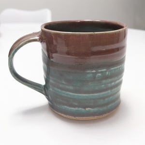 May include: A handmade ceramic mug with a brown top and a teal-green lower section. The mug has a curved handle and a textured, ridged design. The mug is suitable for hot or cold beverages.
