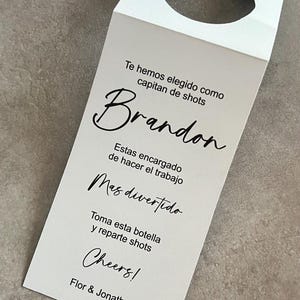 Op de afbeelding: Een wit papieren fleslabel met een cirkelvormig gat aan de bovenkant. Het label heeft een zwart script met de tekst "Te hemos elegido como capitan de shots Brandon Estas encargado de hacer el trabajo Mas divertido Toma esta botella y reparte shots Cheers! Flor & Jonathan."