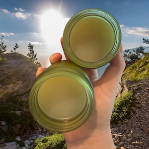 May include: Two open, green-tinted glass jars held up against a bright, sunny sky. The jars are filled with a light-colored substance. The background features a mountain landscape with trees and rocks.