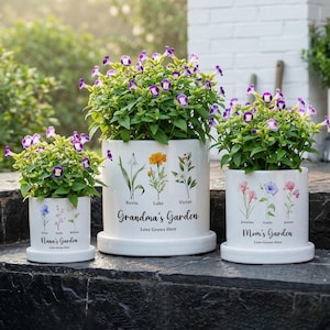 May include: Three white ceramic flower pots with floral designs and text. The pots are filled with green plants and purple and white flowers. The text on the pots reads "Nana's Garden", "Grandma's Garden", and "Mom's Garden".