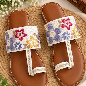May include: A pair of flat sandals with brown footbeds and white straps. The wide top strap features intricate floral embroidery in shades of purple, pink, and yellow. A thin, braided white strap connects to a toe ring adorned with small, clear crystals.
