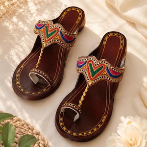 Puede incluir: Un par de sandalias de madera marrón oscuro con diseño de separador de dedos. Las correas superiores están decoradas con bordados vibrantes en hilos rojo, verde, azul y dorado, formando un patrón de corazón. Los bordes tienen costuras doradas.