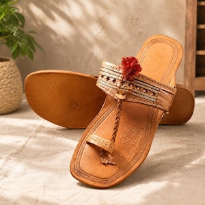 May include: A pair of tan leather sandals featuring a toe post, a braided strap, and a wide horizontal band with decorative stitching and metallic accents. A small red tassel is attached to the center of the band. The footbed is embossed with text.