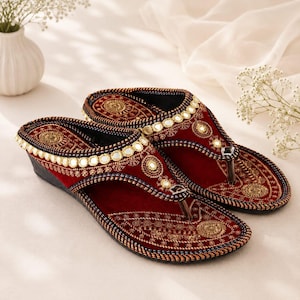 Puede incluir: Un par de sandalias de cuña de terciopelo rojo oscuro con intrincados bordados dorados y adornos de espejos circulares en las tiras y la plantilla. Las sandalias tienen un separador de dedos y un tacón de cuña oscuro con costuras detalladas.