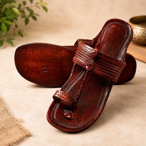 Puede incluir: Un par de sandalias de cuero marrón oscuro con diseño de trabilla para el dedo, detalles trenzados intrincados a lo largo de la tira central y una banda horizontal ancha en la parte superior. El calzado tiene suela plana y acabado texturizado.