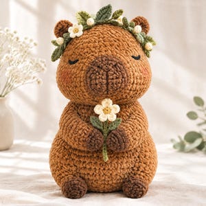 May include: A brown crocheted capybara plush toy featuring a floral crown made of green leaves and small white flowers. The capybara has closed eyes, pink cheeks, and is holding a single white flower with a yellow center in its front paws.