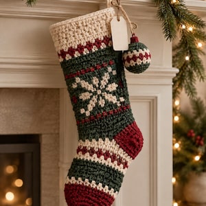 May include: A crocheted Christmas stocking featuring a cream-coloured cuff, a dark green body with a white snowflake pattern, and a deep red heel and toe. A small matching crocheted ornament and a blank rectangular gift tag hang from the top.