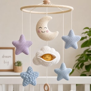 Puede incluir: Un móvil de ganchillo para bebé que presenta una luna creciente de color crema con una cara sonriente, una nube blanca que contiene un pequeño sol amarillo y cuatro estrellas en tonos morado claro y azul, suspendidos de un marco de madera.