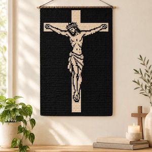 May include: A rectangular wall hanging featuring a crochet design of a figure on a cross. The piece uses a high-contrast color scheme with a solid black background and a cream-colored figure and cross. It is suspended from a wooden dowel.