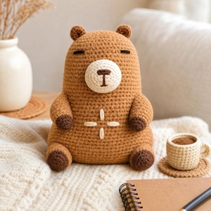 Peut inclure: Une peluche ours crochetée marron avec un museau crème, des pattes marron foncé et des yeux fermés. L'ours présente une broderie en forme de croix sur la poitrine. À côté se trouve une petite tasse crochetée assortie couleur crème sur un sous-verre.