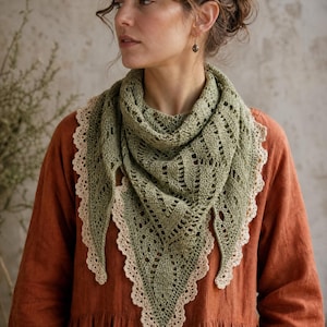 May include: A triangular crochet shawl in sage green featuring an intricate openwork pattern and a scalloped cream-coloured lace border. The shawl is draped around the neck and shoulders, showcasing the detailed yarn texture and craftsmanship.