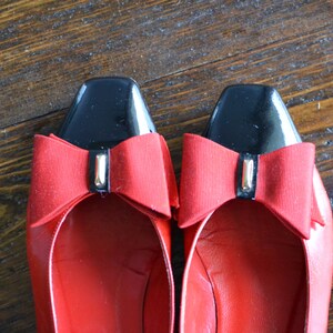 May include: A pair of red leather flats with black square toes. Each shoe features a red bow with a gold-colored accent. The shoes are displayed on a wooden surface.