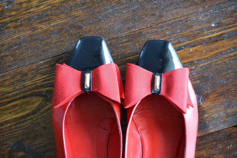 May include: A pair of red leather flats with black square toes. Each shoe features a red bow with a gold-colored accent. The shoes are displayed on a wooden surface.
