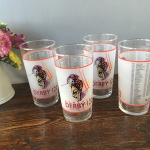 May include: Four clear drinking glasses with a frosted white band featuring a horse racing graphic and the text "Kentucky Derby 122". The glasses have a list of names and dates on the side.