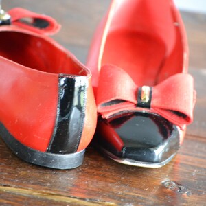 May include: A pair of red flats with black patent leather accents. Each shoe features a large red bow with a decorative embellishment. The shoes are displayed on a wooden surface.