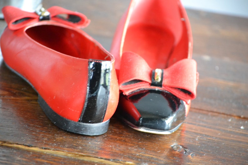 May include: A pair of red flats with black patent leather accents. Each shoe features a large red bow with a decorative embellishment. The shoes are displayed on a wooden surface.