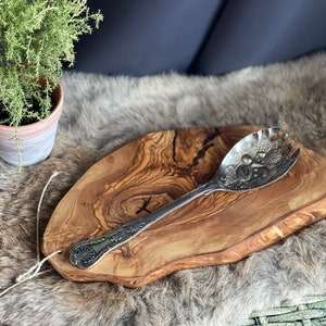 May include: A silver serving spoon with a decorative design sits on a large, rustic olive wood serving board. The board has a natural, irregular shape and a light brown colour.