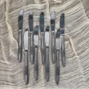 May include: A set of ten silver dinner knives with ornate handles. The knives are laid out on a beige fabric background.