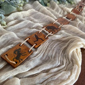 Puede incluir: Una guirnalda de madera con imágenes de personas y animales, conectadas por un cordel. La guirnalda está sobre un fondo de tela blanca.