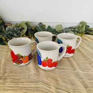 Pode incluir: Conjunto de quatro canecas de cerâmica branca com ilustrações coloridas de frutas. Cada caneca apresenta um design de fruta diferente, incluindo morangos, tomates e pimentões. As canecas têm uma forma simples e arredondada e uma alça no lado.