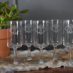 Può includere: Un set di sei flûte da champagne trasparenti. Ogni bicchiere presenta un disegno inciso di una figura all'interno di un rombo. I bicchieri sono esposti su una superficie simile alla pelliccia, con una pianta in vaso sullo sfondo.