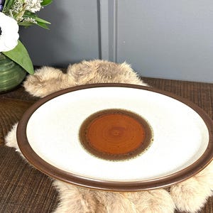 May include: An oval-shaped ceramic serving platter with a white speckled center and a brown border. The center features a concentric circle design in shades of brown. A decorative piece for serving food.