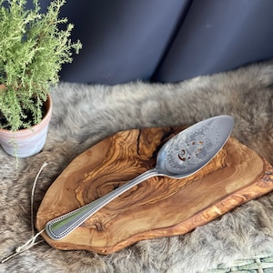 May include: A silver cake server with an ornate design sits on a wooden chopping board. The board has a natural wood grain and is made of olive wood.