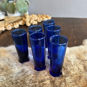 May include: Six cobalt blue shot glasses with a swirl design. The glasses are arranged in a semi-circle on a brown fur surface.