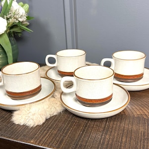 May include: Four matching coffee cups and saucers. The cups are white with a speckled texture and a brown band around the middle. The saucers have a matching brown trim. The set is on a wooden surface.