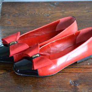 May include: A pair of red leather flats with black patent leather toe caps. Each shoe features a red bow with a gold-colored metal accent. The shoes are displayed on a wooden surface.
