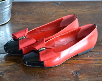 May include: A pair of red leather flats with black patent leather toe caps. Each shoe has a red bow with a gold-coloured metal accent. The shoes are displayed on a wooden surface.