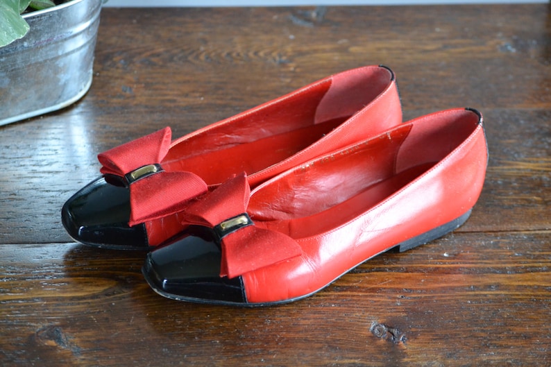 May include: A pair of red leather flats with black patent leather toe caps. Each shoe features a red bow with a gold-colored metal accent. The shoes are displayed on a wooden surface.