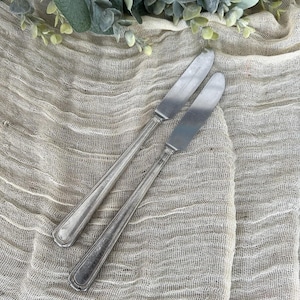 Op de afbeelding: Twee zilveren botermessen met afgeronde bladen en gedetailleerde handvatten liggen op een getextureerde, beige doek. De messen staan schuin naar elkaar toe. Groene en witte bladeren accentueren de bovenkant van de afbeelding.