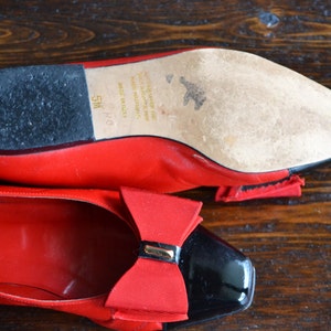 May include: Red leather ballet flats with black patent leather toe caps and a red bow detail. The sole is visible, showing wear and the size 5M. The shoes are on a wooden surface.
